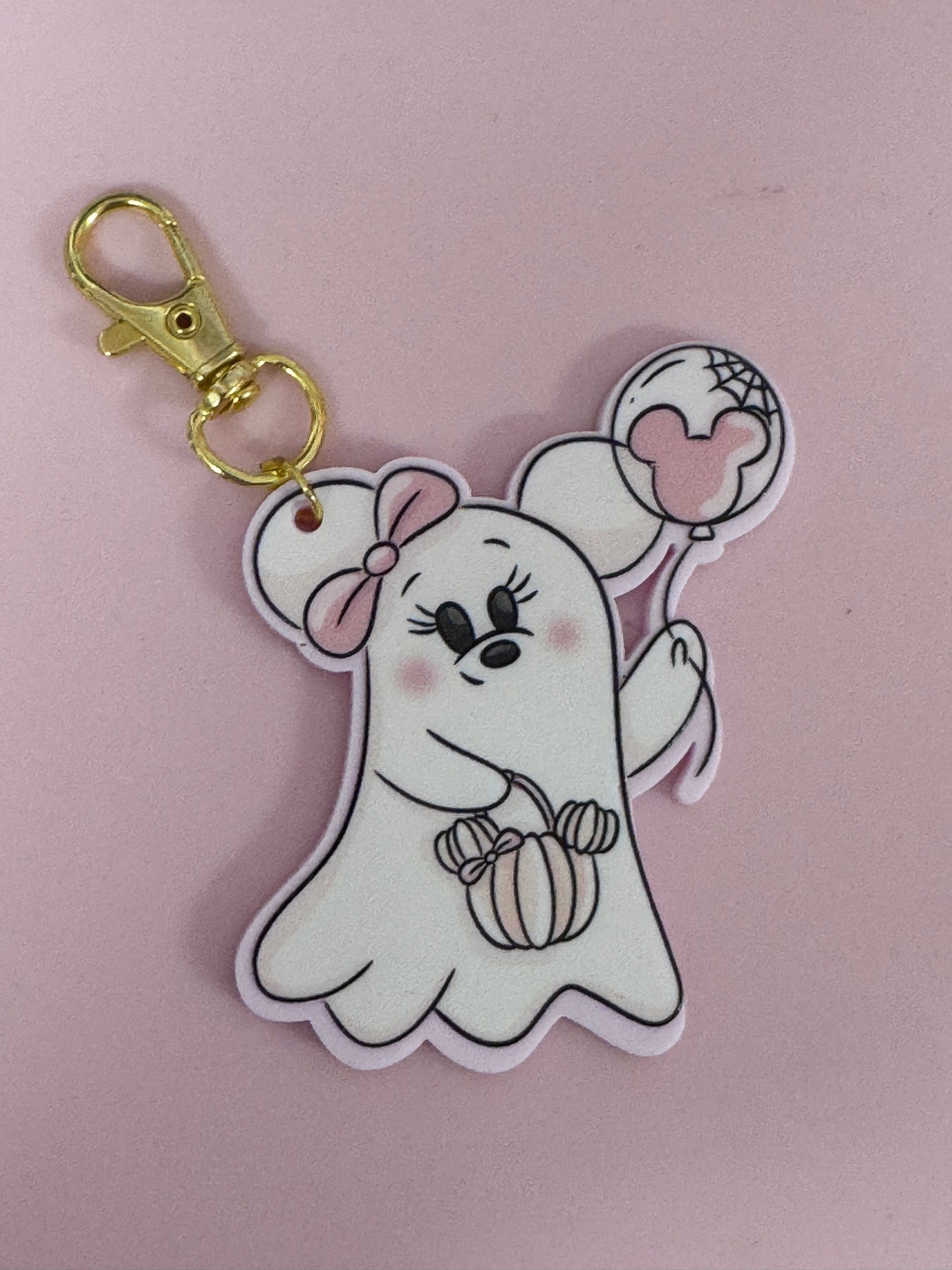 A cute ghost-shaped keychain charm featuring a white cartoon ghost with a pink bow, holding a striped pumpkin and a pink heart-shaped balloon, attached to a gold clasp on a solid pink background.